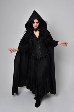Siyah Viktorya dönemi cadı kostümü giymiş, pelerinli koyu renk saçlı bir kadının tam boy portresi. Stüdyonun arka planında, hareketli el hareketleri ile ayakta duruyoruz..