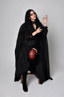 Siyah Viktorya dönemi cadı kostümü giymiş, pelerinli koyu renk saçlı bir kadının tam boy portresi. Stüdyonun arka planında, hareketli el hareketleri ile ayakta duruyoruz..