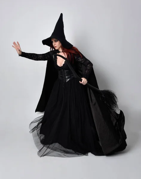 身穿黑色维多利亚式女巫服装的黑头发女人的全长肖像 站立姿势 手持带有手势动作的扫帚棒 在工作室背景下 — 图库照片
