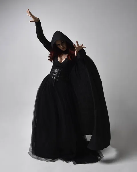 全长的画像 深色头发的女人穿着黑色维多利亚式的女巫服装和一件飘逸的斗篷 站立姿势 有手势的手部动作 在工作室背景下 — 图库照片