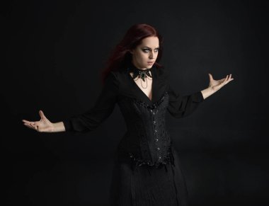 Siyah Victoria Cadı kostümü giyen kızıl saçlı kadının yakın plan portresi. Duruş, hareketli el hareketleri, karanlık stüdyo arka planı.