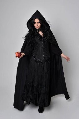 Siyah Victoria tarzı cadı kostümü giyen, stüdyo arka planında el hareketleriyle duran koyu renk saçlı bir kadının tam boy portresi..