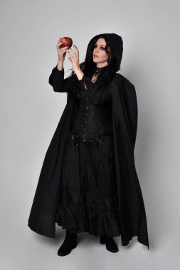 Siyah Victoria tarzı cadı kostümü giyen, stüdyo arka planında el hareketleriyle duran koyu renk saçlı bir kadının tam boy portresi..