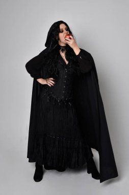 Siyah Victoria tarzı cadı kostümü giyen, stüdyo arka planında el hareketleriyle duran koyu renk saçlı bir kadının tam boy portresi..