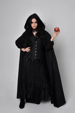 Full length portrait of dark haired woman wearing  black victorian witch costume  standing pose, with  gestural hand movements,  against studio background. clipart