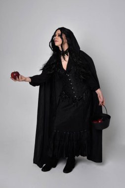 Siyah Victoria tarzı cadı kostümü giyen, stüdyo arka planında el hareketleriyle duran koyu renk saçlı bir kadının tam boy portresi..