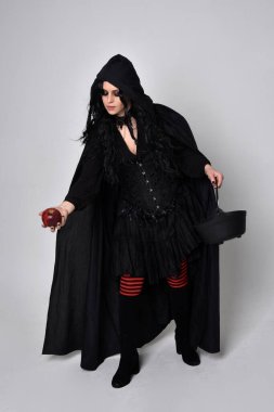 Siyah Victoria tarzı cadı kostümü giyen, stüdyo arka planında el hareketleriyle duran koyu renk saçlı bir kadının tam boy portresi..