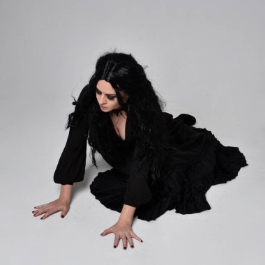 Siyah Victoria tarzı cadı kostümü giyen, stüdyo arka planında hareketli, hareketli, siyah saçlı bir kadının tam boy portresi..