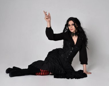 Siyah Victoria tarzı cadı kostümü giyen, stüdyo arka planında hareketli, hareketli, siyah saçlı bir kadının tam boy portresi..