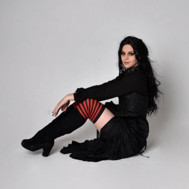 Siyah Victoria tarzı cadı kostümü giyen, stüdyo arka planında hareketli, hareketli, siyah saçlı bir kadının tam boy portresi..