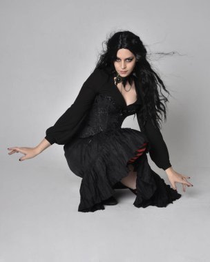 Siyah Victoria tarzı cadı kostümü giyen, stüdyo arka planında hareketli, hareketli, siyah saçlı bir kadının tam boy portresi..