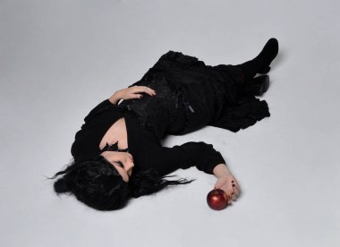 Siyah Victoria tarzı cadı kostümü giyen, yerde yatan, ölü gibi uyuyan, stüdyo arka planında soyutlanmış koyu renk saçlı kadının tam boy portresi..