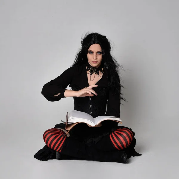 Full length portrait of dark haired woman wearing  black victorian witch costume  sitting pose, with  gestural hand movements,  against studio background.