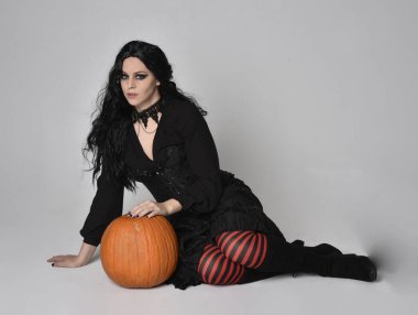 Siyah Viktorya dönemi cadı kostümü giymiş uzun saçlı bir kadının uzun portresi stüdyo arka planında el hareketleriyle balkabağı pozu veriyor..