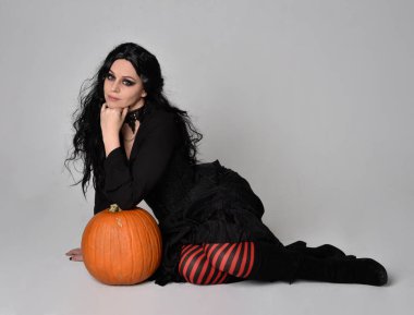 Siyah Viktorya dönemi cadı kostümü giymiş uzun saçlı bir kadının uzun portresi stüdyo arka planında el hareketleriyle balkabağı pozu veriyor..