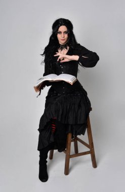 Siyah Victoria cadısı kostümü giymiş koyu renk saçlı bir kadının tam boy portresi. Sandalyede otururken, el hareketleriyle, stüdyo arka planında..