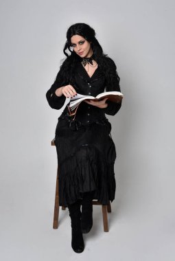 Siyah Victoria cadısı kostümü giymiş koyu renk saçlı bir kadının tam boy portresi. Sandalyede otururken, el hareketleriyle, stüdyo arka planında..