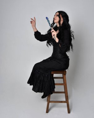 Siyah Victoria cadısı kostümü giymiş koyu renk saçlı bir kadının tam boy portresi. Sandalyede otururken, el hareketleriyle, stüdyo arka planında..