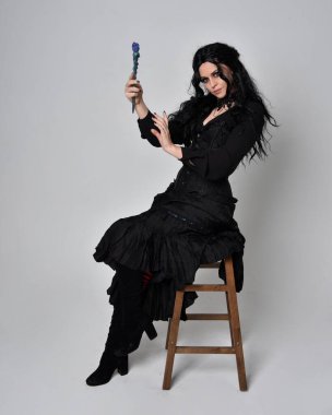 Siyah Victoria cadısı kostümü giymiş koyu renk saçlı bir kadının tam boy portresi. Sandalyede otururken, el hareketleriyle, stüdyo arka planında..
