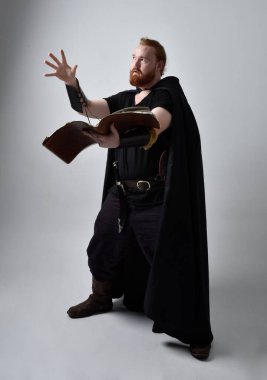 Ortaçağ Viking 'i giyen kızıl saçlı adamın portresi pelerinli fantezi kostümüne ilham verdi. Elinde sihirli bir kitapla ayakta duruyor, el hareketleriyle sanki bir büyü yapıyor gibi, stüdyo arka planında izole edilmiş..