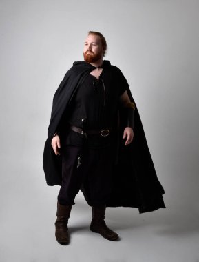  Ortaçağ Viking 'i giyen kızıl saçlı adamın portresi koyu renk pelerinli fantezi kostümüne ilham verdi. Stüdyo arka planına izole edilmiş bir büyü yapar gibi hareketli poz veriyor..