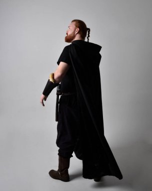  Ortaçağ Viking 'i giyen kızıl saçlı adamın portresi koyu renk pelerinli fantezi kostümüne ilham verdi. Stüdyo arka planına izole edilmiş bir büyü yapar gibi hareketli poz veriyor..