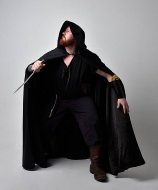  Ortaçağ Viking 'i giyen kızıl saçlı adamın portresi koyu renk pelerinli fantezi kostümüne ilham verdi. Stüdyo arka planına izole edilmiş bir büyü yapar gibi hareketli poz veriyor..