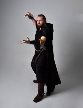  Ortaçağ Viking 'i giyen kızıl saçlı adamın portresi koyu renk pelerinli fantezi kostümüne ilham verdi. El hareketleriyle poz vermek, sanki büyülü bir büyü yapmak gibi, stüdyo arka planında izole edilmiş..