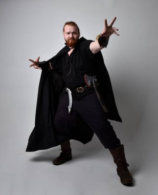  Ortaçağ Viking 'i giyen kızıl saçlı adamın portresi koyu renk pelerinli fantezi kostümüne ilham verdi. El hareketleriyle poz vermek, sanki büyülü bir büyü yapmak gibi, stüdyo arka planında izole edilmiş..