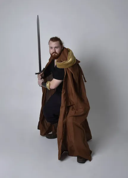 portrait of red haired man wearing medieval viking inspired fantasy costume, holding a long sword weapon. Isolated against studio background