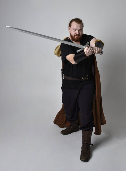  portrait of red haired man wearing medieval viking inspired fantasy costume, holding a long sword weapon. Isolated against studio background
