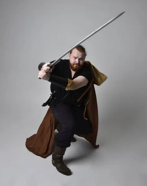 中世のバイキングを着た赤い髪の男の肖像画は 長い剣の武器を保持し ファンタジーの衣装にインスピレーションを与えた スタジオの背景に隔離された — ストック写真
