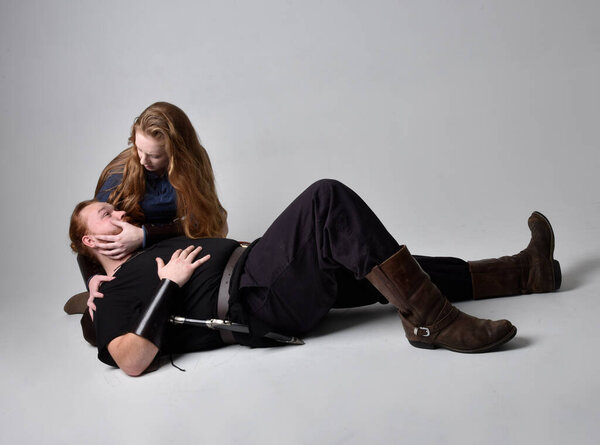 Full length  portrait of red haired  couple, man and woman wearing medieval viking inspired fantasy costumes, sitting on ground as if wounded from battle injuries in combat, isolated on white  studio background.
