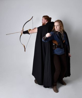 Kızıl saçlı bir çiftin tam boy portresi, ortaçağ Viking ilham verici fantezi kostümleri giyen bir kadın ve erkek, elinde okçuluk yayı, ok ve uzun kılıç silahları olan, beyaz stüdyo arka planında izole edilmiş bir şekilde ayakta duran bir dövüş pozisyonu..