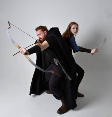 Kızıl saçlı bir çiftin tam boy portresi, ortaçağ Viking ilham verici fantezi kostümleri giyen bir kadın ve erkek, elinde okçuluk yayı, ok ve uzun kılıç silahları olan, beyaz stüdyo arka planında izole edilmiş bir şekilde ayakta duran bir dövüş pozisyonu..