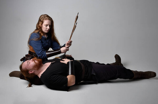 Full length  portrait of red haired  couple, man and woman wearing medieval viking inspired fantasy costumes, sitting on ground as if wounded from battle injuries in combat, isolated on white  studio background.