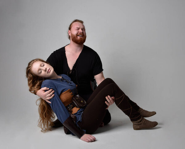 Full length  portrait of red haired  couple, man and woman wearing medieval viking inspired fantasy costumes, sitting on ground as if wounded from battle injuries in combat, isolated on white  studio background.