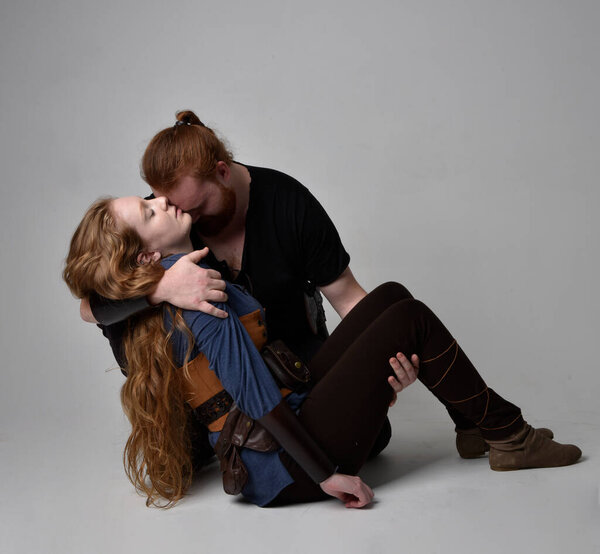 Full length  portrait of red haired  couple, man and woman wearing medieval viking inspired fantasy costumes, sitting on ground as if wounded from battle injuries in combat, isolated on white  studio background.