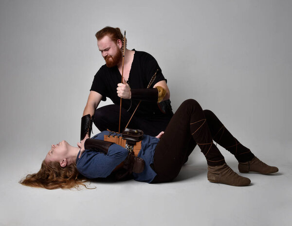 Full length  portrait of red haired  couple, man and woman wearing medieval viking inspired fantasy costumes, sitting on ground as if wounded from battle injuries in combat, isolated on white  studio background.