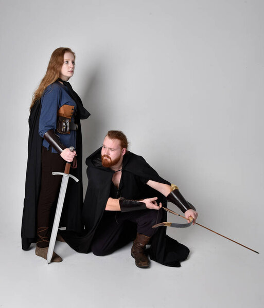 Full length  portrait of red haired  couple, man and woman wearing medieval viking inspired fantasy costumes, standing fighting pose holding  archery bow and arrow and long sword weapons, isolated on white  studio background.