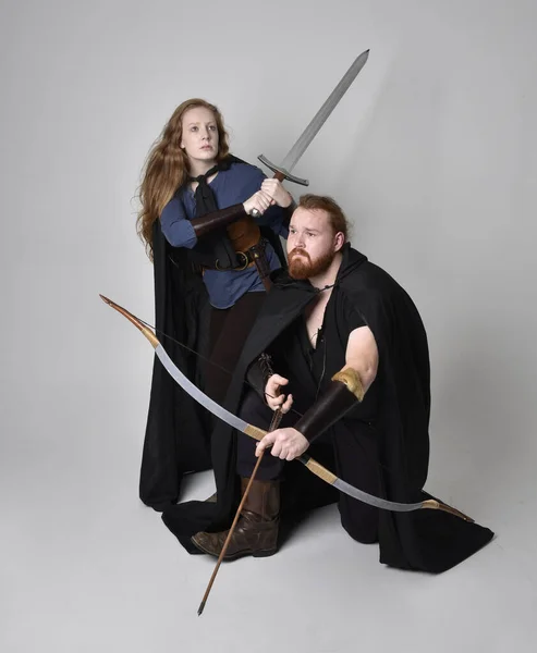 Full length  portrait of red haired  couple, man and woman wearing medieval viking inspired fantasy costumes, standing fighting pose holding  archery bow and arrow and long sword weapons, isolated on white  studio background.