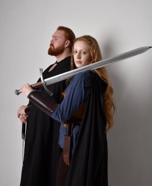 Kızıl saçlı bir çiftin tam boy portresi, ortaçağ Viking 'i giymiş bir adam ve kadın ilham verici fantezi kostümleri, duruş pozisyonu kılıç tutuş, beyaz stüdyo arka planında izole edilmiş.