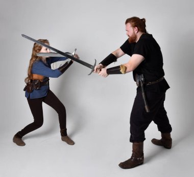 Kızıl saçlı bir çiftin tam boy portresi, ortaçağ Viking 'i giymiş bir adam ve kadın ilham verici fantezi kostümleri, duruş pozisyonu kılıç tutuş, beyaz stüdyo arka planında izole edilmiş.