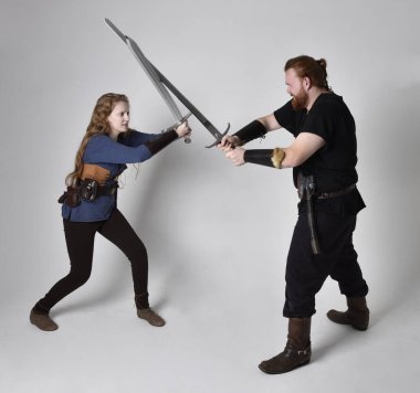Kızıl saçlı bir çiftin tam boy portresi, ortaçağ Viking 'i giymiş bir adam ve kadın ilham verici fantezi kostümleri, duruş pozisyonu kılıç tutuş, beyaz stüdyo arka planında izole edilmiş.