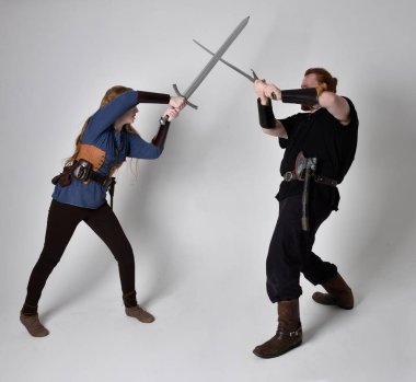 Kızıl saçlı bir çiftin tam boy portresi, ortaçağ Viking 'i giymiş bir adam ve kadın ilham verici fantezi kostümleri, duruş pozisyonu kılıç tutuş, beyaz stüdyo arka planında izole edilmiş.