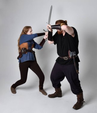 Kızıl saçlı bir çiftin tam boy portresi, ortaçağ Viking 'i giymiş bir adam ve kadın ilham verici fantezi kostümleri, duruş pozisyonu kılıç tutuş, beyaz stüdyo arka planında izole edilmiş.