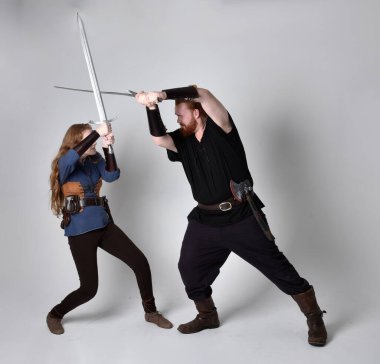 Kızıl saçlı bir çiftin tam boy portresi, ortaçağ Viking 'i giymiş bir adam ve kadın ilham verici fantezi kostümleri, duruş pozisyonu kılıç tutuş, beyaz stüdyo arka planında izole edilmiş.