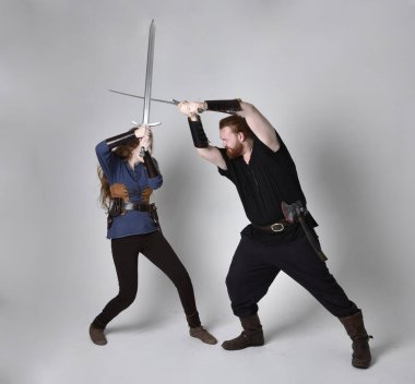 Kızıl saçlı bir çiftin tam boy portresi, ortaçağ Viking 'i giymiş bir adam ve kadın ilham verici fantezi kostümleri, duruş pozisyonu kılıç tutuş, beyaz stüdyo arka planında izole edilmiş.