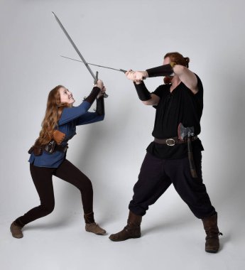 Kızıl saçlı bir çiftin tam boy portresi, ortaçağ Viking 'i giymiş bir adam ve kadın ilham verici fantezi kostümleri, duruş pozisyonu kılıç tutuş, beyaz stüdyo arka planında izole edilmiş.