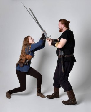 Kızıl saçlı bir çiftin tam boy portresi, ortaçağ Viking 'i giymiş bir adam ve kadın ilham verici fantezi kostümleri, duruş pozisyonu kılıç tutuş, beyaz stüdyo arka planında izole edilmiş.